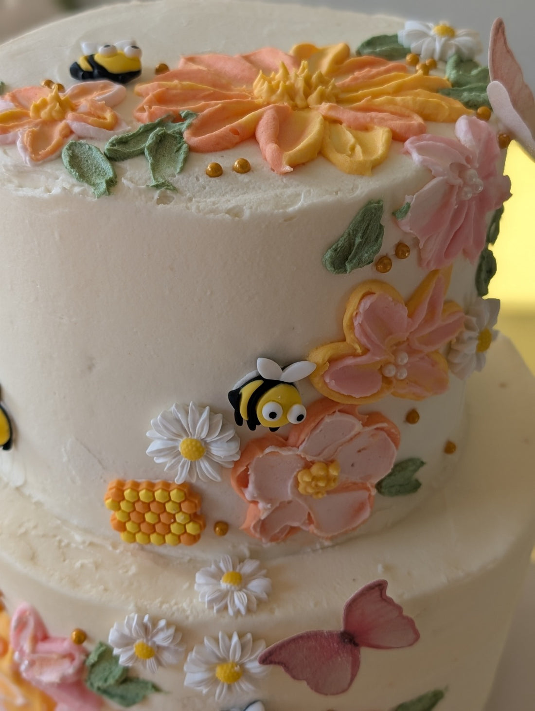 Two-tier buttercream cake with pink and orange flowers, bees, daisies, and honeycomb details from Happy Hippo Bakery in Hutto, Texas